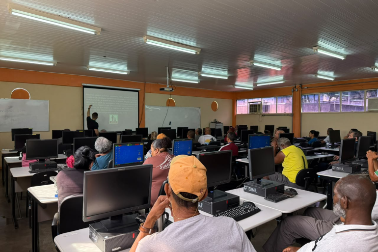 Sala de aula equipada com computadores. Professor em frente ao quadro e alunos sentados de frente para computadores, assistindo a aula.