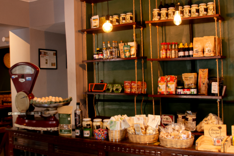 Produtos como biscoitos, café, temperos e outros itens da culinária mineira estão dispostos em prateleiras no Armazém Roça Grande. Na imagem, além dos produtos, está um modelo de balança antiga.
