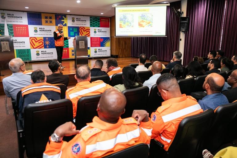 Imagem mostra várias pessoas uniformizadas com coletes laranja assistindo a uma apresentação de um homem tambémn de colete laranja. No fundo há um banner com logomarcas da PBH.