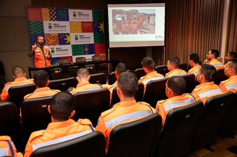 Imagem mostra uma pessoa da Defesa Civil, vestoda com uma colete laranja em uma cenário com uma apresentação de power point atrás dele. Há também uma banner com identidade visual da Prefeitura. Na platéia há vários oficiais dos Bombeiros assistindo à palestra. Todos estão com uniformes laranja do coorporação.