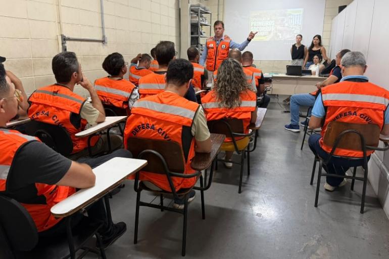 Defesa Civil de BH capacita agentes em Primeiros Socorros Psicológicos para atendimento em situações de crise vários agentes da Defesa Civil assistem a uma palestra. Todos estão com coletes laranja, exceto três pessoas ao fundo.