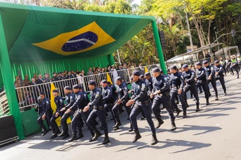 Posto de Comando será aberto no COP-BH para monitoramento do desfile de 7 de setembro na Afonso Pena Guardas Municipais marchando em formação em frente a um palco coberto com uma grande bandeira brasileira pendurada atrás deles. O palco estão autoridades e oficiais.