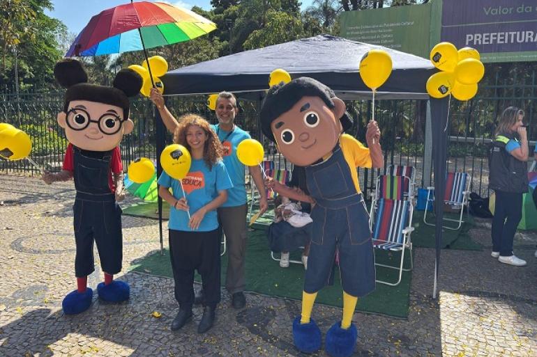 Imagem mostra uma criança e um adulto ladeados por dois mascotes em um estande da campanha.