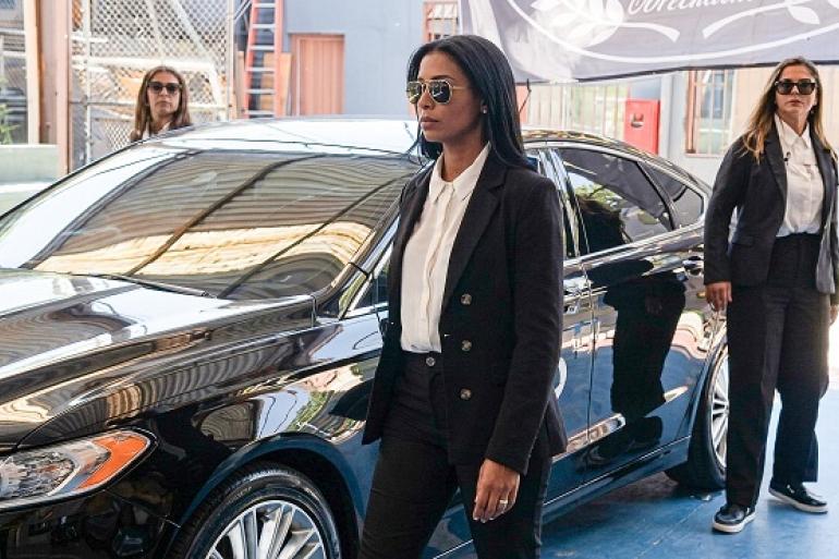 Uma mulher caminha usando óculos escuros, camisa branca e terno preto. Ao lado dela, há um carro estacionado em uma área coberta. Ao fundo e à direita, outras duas mulheres vestidas com trajes semelhantes e óculos escuros mantêm uma postura atenta.