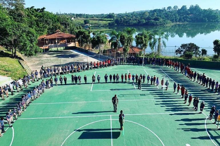Escotismo da Guarda Municipal completa 20 anos com a participação de mais de 500 crianças e adolescentes de BH Grupo de jovens e adultos do projeto de escotismo vestindo uniformes são organizados em formato de "U" sobre uma quadra poliesportiva ao ar livre. Ao fundo, destaca-se uma paisagem natural, com um lago sob um céu azul sem nuvens.