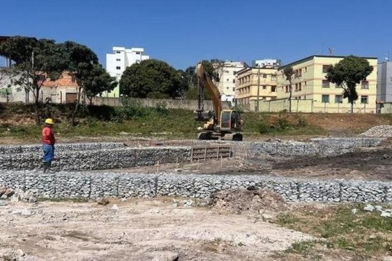 Imagem de um funcionário, com camisa vermelha, trabalhando na obra de um projeto piloto do alagado na Bacia Várzea da Palma, com um trator e prédios ao fundo. 