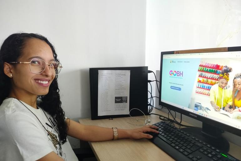 PBH oferece 58 vagas de estágio em setembro com bolsas de até R$ 1,8 mil Imagem de uma jovem de cabelo longo, óculos, sorrindo, mexendo no teclado de um computador à sua frente, em um ambiente com paredes brancas.