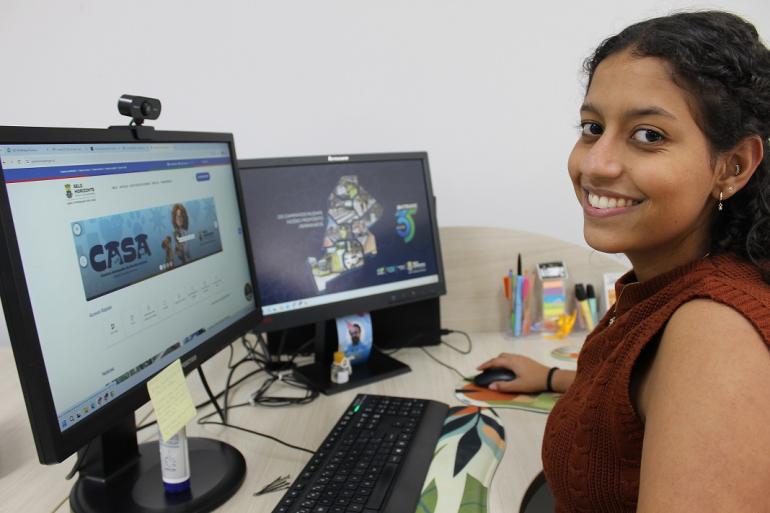 Prefeitura de Belo Horizonte oferece 69 vagas de estágio em outubro com bolsa de até R$ 1.886,59 Descrição da imagem: Uma jovem está sentada em frente a dois computadores, trabalhando em uma mesa de escritório. Ela aparece sorrindo para a câmera, enquanto utiliza o mouse. Em um dos monitores, é possível ver uma página relacionada à Prefeitura de Belo Horizonte. Sobre a mesa há teclado, mouse, materiais de escritório e recipientes com canetas.