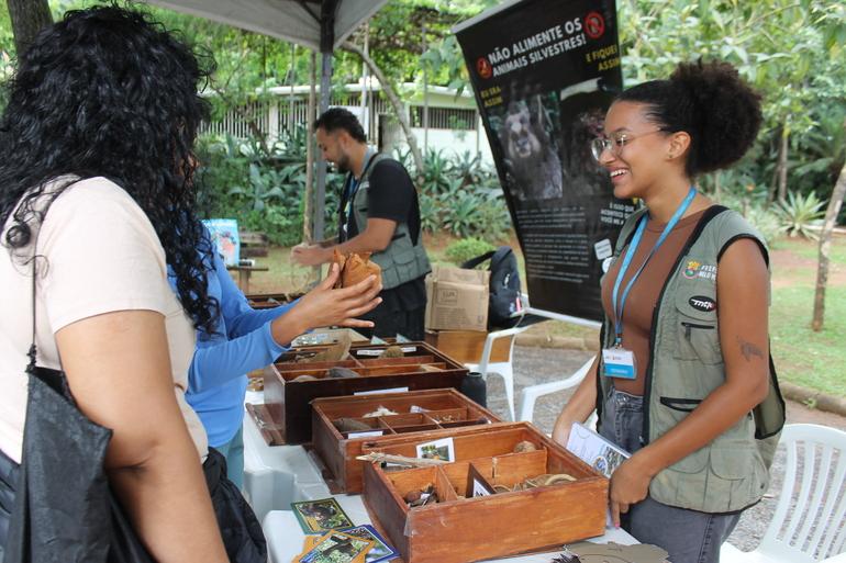 É tempo de férias com programação especial nos parques e Zoobotânica Mulher usando colete da Prefeitura de Belo Horizonte interagindo com visitantes do parque, onde caixas com sementes e materiais naturais estão expostos, ao lado de banner educativo sobre não alimentar animais silvestres