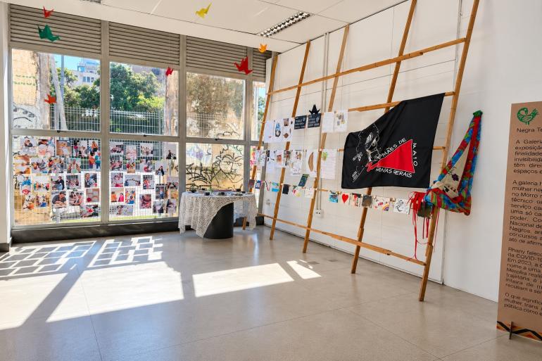  Fotografia de uma galeria de exposição iluminada por grandes janelas. Fotografias, desenhos e cartazes estão expostos em estruturas de madeira. Ao centro, há uma mesa com objetos. À direita, destaca-se uma bandeira preta e vermelha da Marcha das Mulheres de Minas Gerais. No teto, pequenas dobraduras de papel coloridas estão suspensas.