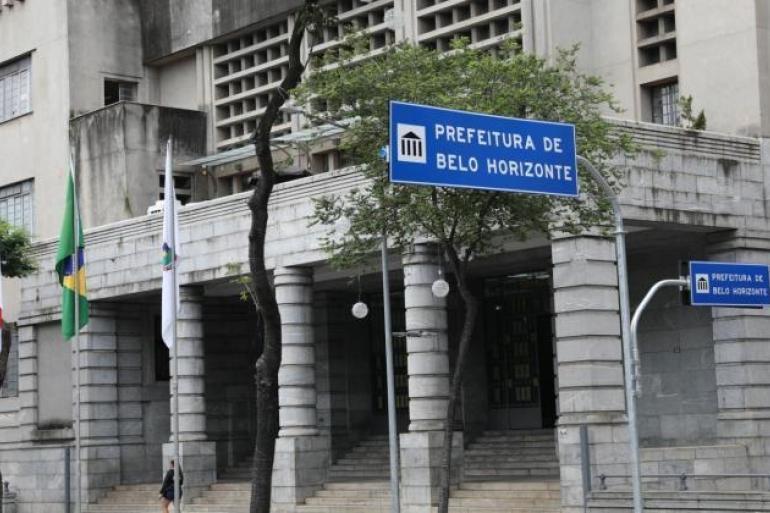 Uso de IA na análise de Notas Fiscais de Serviços Eletrônicos coloca PBH como finalista do Concurso Inovação no Setor Público A imagem mostra a fachada do prédio sede da Prefeitura de Belo Horizonte, com duas placas onde está escrito Prefeitura de Belo Horizonte, mais uma bandeira do Brasil e uma bandeira de Belo Horizonte hasteadas; na calçada uma pedestre caminha em frente ao prédio.