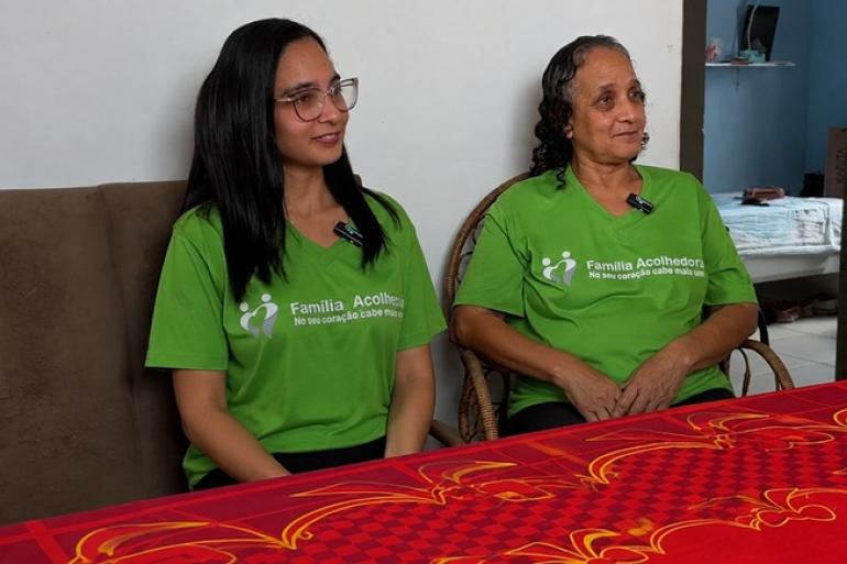 Duas mulheres estão sentadas lado a lado em uma sala, diante de uma mesa coberta por uma toalha vermelha estampada. As duas usam camisetas verdes com a identificação “Família Acolhedora” e estão com microfones de lapela presos à roupa.