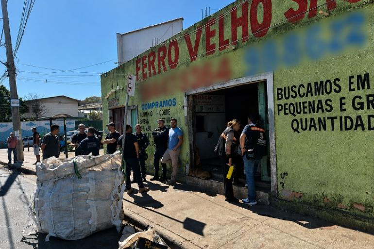 Imagem mo0stra vários fiscais na porta de um estabelecimento que comercializa ferro-velho. Além dos fiscais há várias pessoas e um grande saco branco.