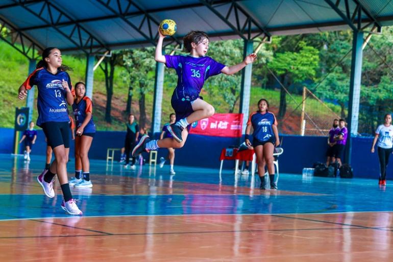 Chamamento de interessados em patrocinar eventos esportivos em BH é publicado pela Prefeitura Em uma quadra coberta de handebol, uma jovem atleta de uniforme roxo, número 36, salta com a bola erguida na mão direita, preparando o arremesso. Ao redor, jogadoras da equipe adversária, de uniforme azul-marinho com detalhes laranja, acompanham a jogada. Ao fundo, aparecem outras atletas, integrantes das equipes e a área externa arborizada.
