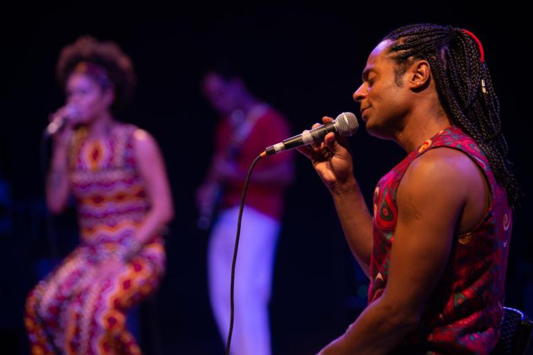 Sonoridades afro-brasileiras marcam show no Teatro Raul Belém Machado Fotografia em que o cantor Ton Guimarães canta com os olhos fechados, segurando o microfone em um palco escuro. Ele tem tranças longas e veste um colete estampado de vermelho. Ao fundo, fora de foco, uma mulher com um macacão colorido também canta e um homem toca violão.