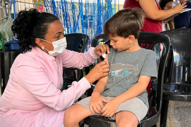 Prefeitura de BH encerra Campanha de Multivacinação com cerca de 73 mil doses aplicadas Criança sentada recebendo uma dose de vacina aplicada durante a Campanha de Multivacinação. Uma profissional de saúde, de máscara e jaleco, aplica a vacina. Cena de vacinação ao ar livre.