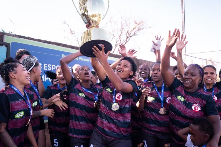 Final do Feminino Adulto é destaque da Copa Centenário neste domingo (30) Fotografia em ambiente externo mostra o time Prointer, campeão da Copa Centenário Feminino Adulto em 2025, reunido em comemoração. No centro, uma jogadora ergue um troféu dourado e prateado acima da cabeça. Ao redor, as companheiras, com uniformes em preto e vinho e medalhas no pescoço, sorriem, gritam e levantam os braços em celebração. Algumas olham para o troféu e outras para as colegas. Ao fundo, há uma placa azul e parte da estrutura do campo.