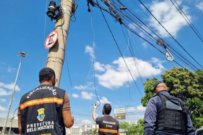 Mutirão da PBH, Cemig e empresas de telecomunicações vai corrigir irregularidades em fios aéreos Foto mostra três agentes da Prefeitura de Belo Horizonte em uma via da cidade, sob um céu azul com nuvens. Dois deles usam coletes pretos com faixas refletivas laranja e a inscrição “Fiscalização – Prefeitura de Belo Horizonte”. O terceiro veste uniforme azul-escuro e colete com a identificação “Guarda Municipal Belo Horizonte”. Os agentes observam e fiscalizam uma grande quantidade de fios e cabos instalados de forma desorganizada em postes. Um dos fiscais, ao centro, segura um cabo que está pendurado.