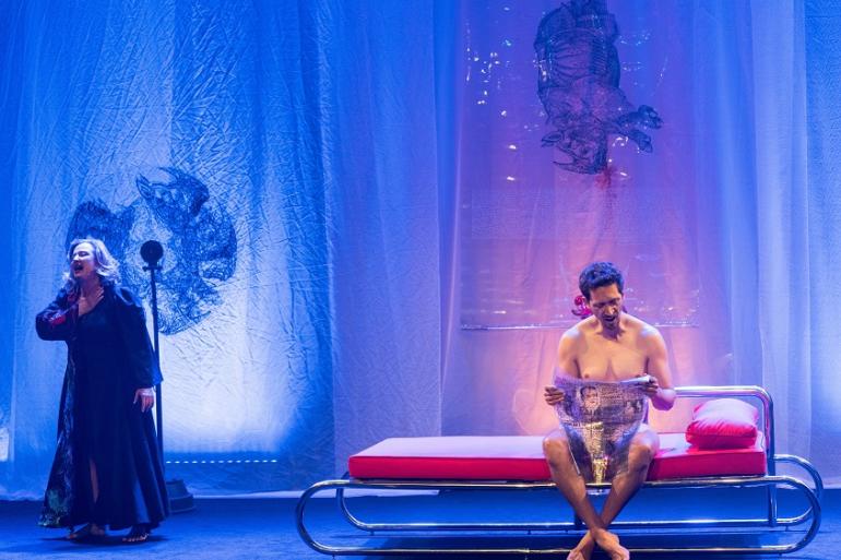 Fotografia de uma cena da peça teatral "O Belo Indiferente". À esquerda, sob uma iluminação azul, uma mulher de vestido longo escuro canta ou gesticula dramaticamente com a mão no peito diante de um microfone pedestal. À direita, num banco/cama de metal com colchão e almofada vermelhos, um homem sem camisa está sentado de pernas cruzadas a ler um jornal. Ao fundo, veem-se cortinas azuis translúcidas com desenhos e ilustrações estampadas.