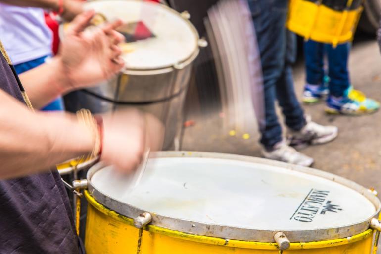Agenda RIC leva samba, cultura popular e ancestralidade a diferentes territórios de BH em agosto Uma imagem em plano médio e close-up foca em um percussionista tocando tambores amarelos. A ação é dinâmica, e suas mãos e as baquetas estão desfocadas para transmitir velocidade e movimento. Ele está segurando uma baqueta no tambor principal em primeiro plano, com o outro tambor e outras pessoas ao fundo.