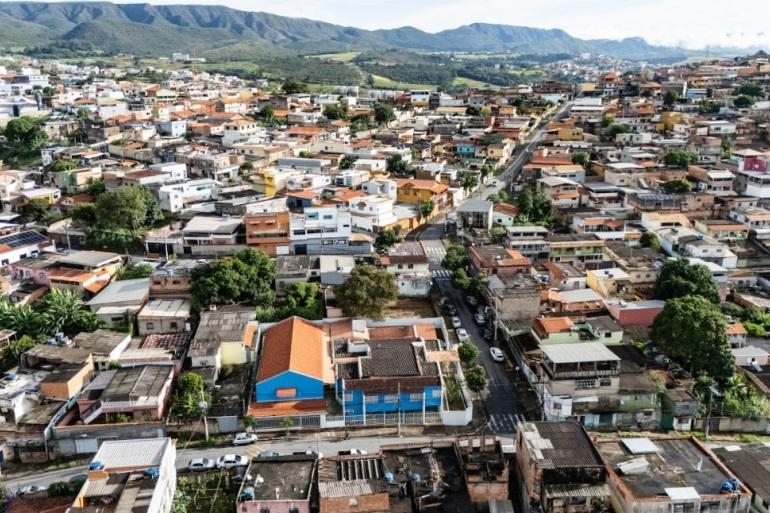 Fotografia aérea de uma área urbana densamente ocupada. Em primeiro plano e ao centro, há diversas casas e pequenos prédios, em sua maioria com telhados de cerâmica ou coberturas de concreto. Uma via asfaltada atravessa o bairro na direção do fundo da imagem, com alguns carros circulando e estacionados.