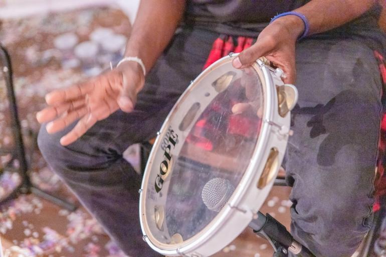 Agenda RIC de setembro reverencia o samba, a cultura negra e as tradições populares Imagem das mãos de um homem negro tocando um pandeiro transparente. A mão direita do homem, que usa uma pulseira branca, está em movimento, posicionada acima do instrumento. Dá para ver que o homem usa uma calça cinza escura.