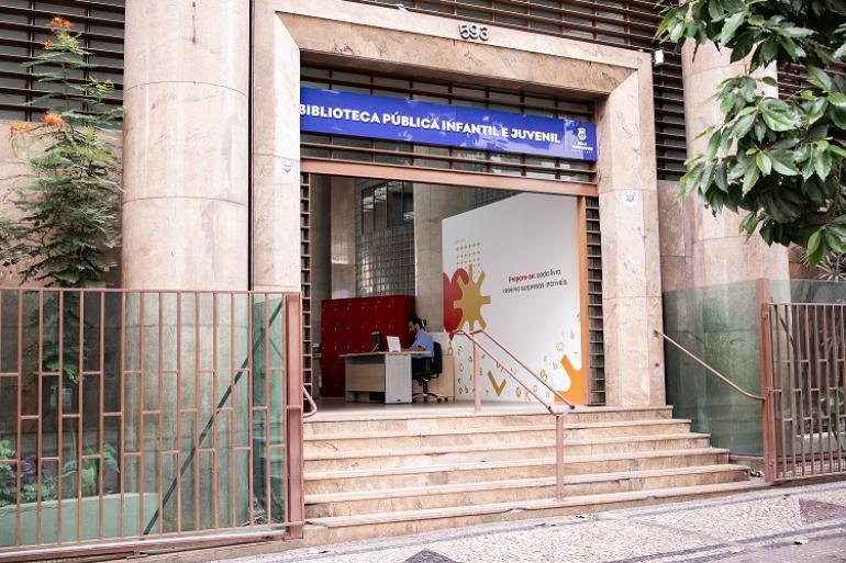 Biblioteca Pública Infantil e Juvenil de BH amplia público e oferta de atividades em um ano na nova sede Imagem mostra a entrada da biblioteca e além do letreiro azul que identifica o local, há também um portal e uma escadaria de mármore. Nas laterais há grades.