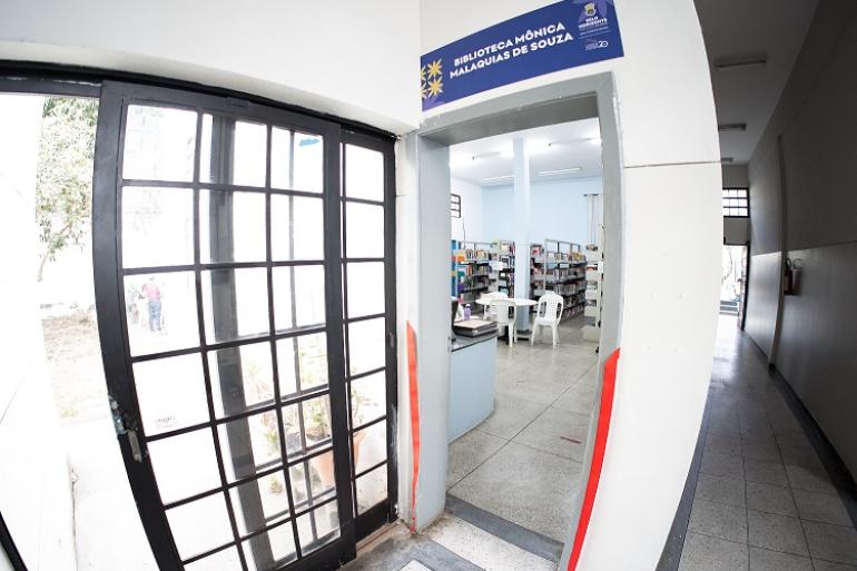 Homenagem do CCSB eterniza legado da bibliotecária Mônica Malaquias Fotografia em ângulo aberto da entrada da biblioteca. Uma porta cinza está aberta. Acima dela, há uma placa azul onde se lê "Biblioteca Mônica Malaquias de Souza". As fitas vermelhas de inauguração estão cortadas e penduradas nas laterais do batente. Pela abertura da porta, vê-se o interior com estantes de livros, mesa e cadeiras. À esquerda, há uma grande janela quadriculada, e à direita, um corredor longo.