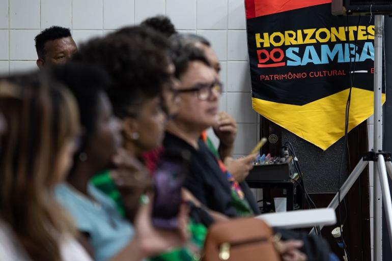 Plano de Salvaguarda do Samba de BH terá terceira etapa de construção coletiva Fotografia colorida de um grupo de pessoas sentadas em fileiras, no canto esquerdo da imagem. Elas estão de perfil, olhando para a direita. As pessoas do primeiro plano estão desfocadas, enquanto o foco principal está em um homem de óculos no centro. No canto superior direito, há um banner vertical pendurado em um tripé de metal, com o design nas cores vermelho, preto e amarelo. No banner, lê-se o título "Horizontes do Samba". A imagem foi registrada durante a 2ª Reunião Pública, em 2025.