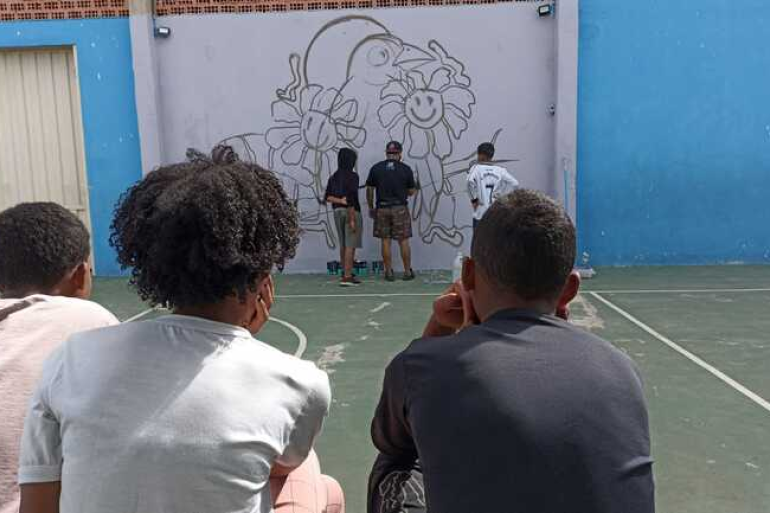 Projeto social da Secretaria de Segurança prevê qualificação profissional e a inclusão produtiva de jovens Três jovens sentados e observando outros jovens fazendo grafitti em um muro branco, a ilustração na parede é um conjunto de flores