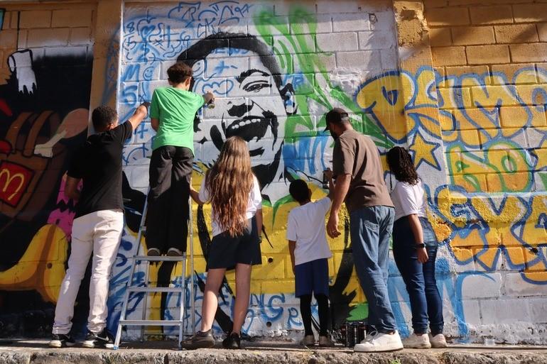 Foto com seis pessoas, três adultos e três crianças, fazendo um grafite com o rosto de uma criança com blusa da seleção brasileira e a frase Rumo ao Hexa