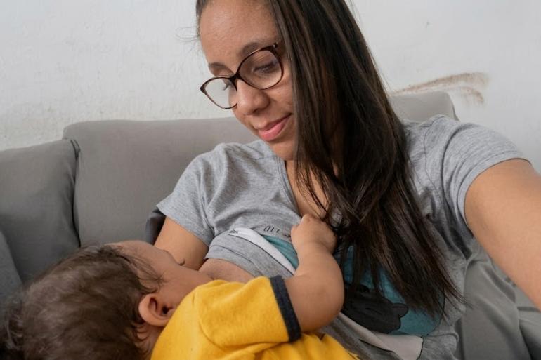 Grupo fortalece apoio à amamentação e promove incentivo à doação de leite Mulher de cabelos longos e escuros, de óculos e camiseta cinza, amamenta um bebê de camiseta amarela. Os dois estão sentados em um sofá. A mulher sorri enquanto olha para o bebê, que mama e segura sua camiseta.