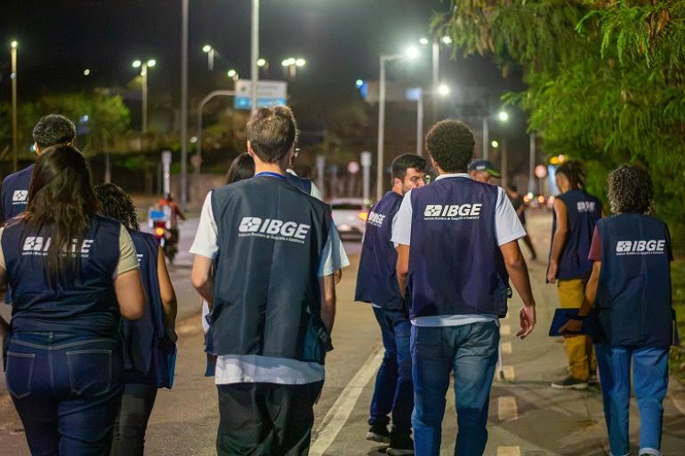  Fotografia noturna feita em uma via pública de Belo Horizonte. Cerca de dez pessoas caminham pela rua, vistas principalmente de costas. Elas usam coletes azul-escuros com o logotipo e a identificação do IBGE
