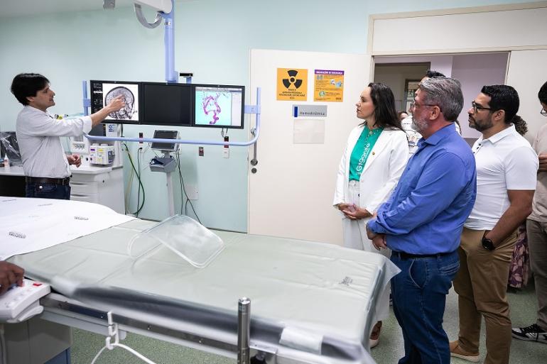 Imagem mostra uma maca em primeiro plano e atrás dela um homem apontando para duas telas de um aparelho do Serviço de Trombectomia Mecânica. O secretário de Saúde, de camisa azul, obserba, ao lado de uma mulher de camisa verde e branca. Há um homem de camiseta branca.