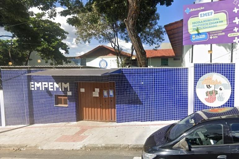 Escolas municipais avançam no Ideb com recomposição das aprendizagens e parceria com as famílias A imagem mostra a fachada da E. M. Edgard da Mata Machado, além de um carro escuro, árvores e casas. O muro da escola é azul e contém a sigla. Na imagem há também uma placa alusiva ao nome da escola.