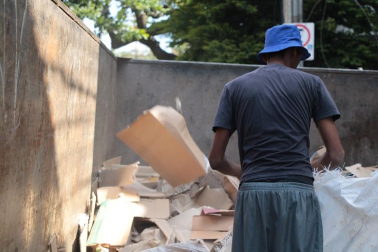 Homem de costas, vestido de camisa cinza e calça verde, com boné azul, está manuseando papelões e materiais recicláveis.