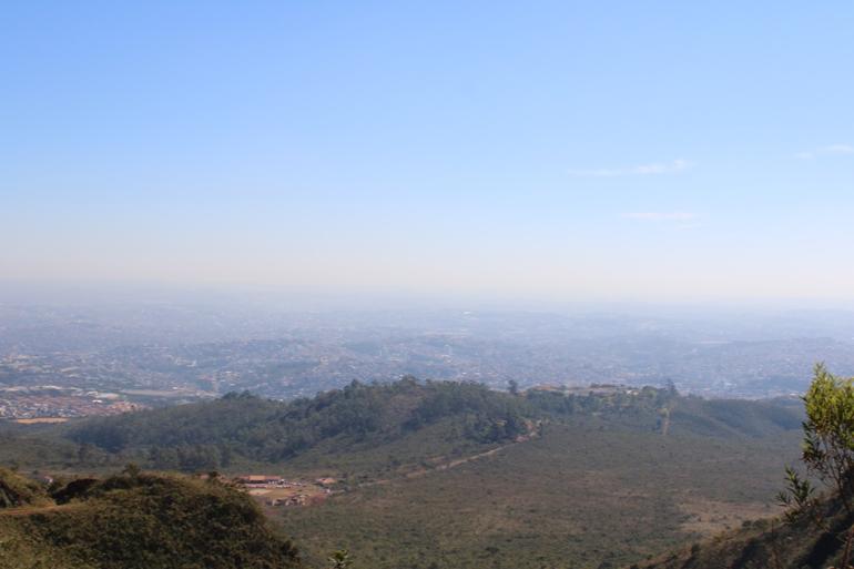 Imagem panorâmica de Belo Horizonte, aérea, com uma parte de vegetação e outra doe uma série de edificações, ao fundo. Dia claro, com uma percepção de névoa de poeira.