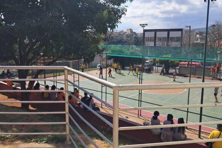Quadra de futebol com alunos jogando. Vários estudantes sentados na arquibancada