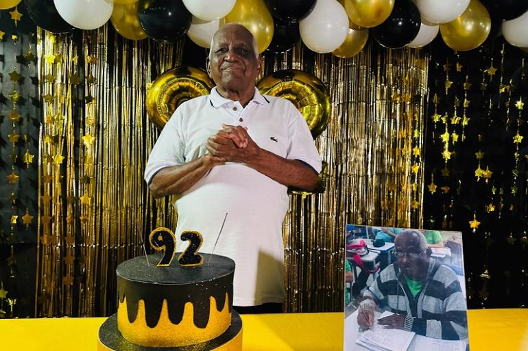 Aluno da EJA comemora aniversário de 92 anos na escola municipal onde mantém rotina de estudos Imagem mostra o senhor José Maria Borges com as mãos postas em frente ao seu bolo de aniversário, que tem uma vela de 92 anos e é dourado e preto, mesmas cores dos bações e da cortina que estão no fundo. Há também uma foto dele em sala de aula.