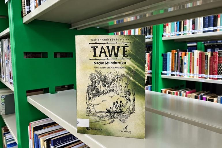 Clube de Leitura do Centro de Educação Ambiental Pampulha promove debate sobre “Tawé – Nação Muduruku” Capa do livro “Tawé – Nação Muduruku”, numa prateleira, tendo como entorno a Ecoteca do Centro de Educação Ambiental da Pampulha.