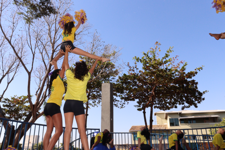 Projeto de solo acrobático traz confiança, sociabilidade e engajamento a estudantes de Venda Nova Quatro líderes de torcida fazendo abertura no chão e uma líder de torcida no alto, fazendo abertura enquanto é carregada por outras duas meninas, todas usando blusa verde amarela do Brasil, em espaço ao ar livre