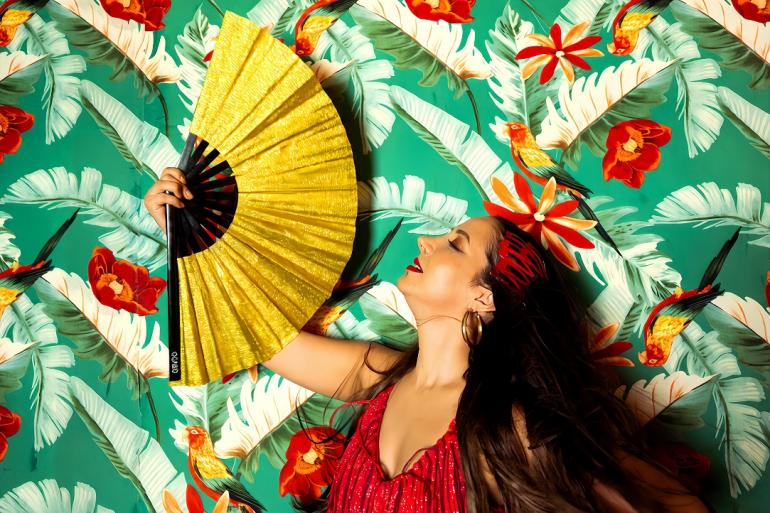 Teatro Raul Belém Machado recebe show autoral da cantora e compositora Lislie neste sábado (19) Cantora vestida de vermelho se abana com um leque e um ambiente decorado com estampa floral vermelha e verde.