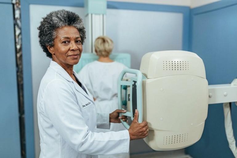 Foto de uma sala de exames de saúde. Em primeiro plano, uma profissional de saúde negra, de cabelos grisalhos e curtos, veste um jaleco branco e segura o equipamento de mamografia. À direita, aparece o aparelho de mamografia, de cor branca, com uma estrutura vertical e uma área de apoio para posicionamento da paciente. Ao fundo, uma mulher, de cabelos claros e usando roupa branca, está de costas diante do equipamento. 