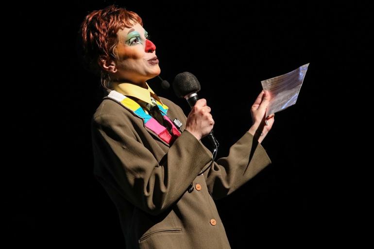 Vagalumes e mudanças climáticas conduzem espetáculo no Teatro Raul Belém Machado A imagem mostra um ator de perfil caracterizada de palhaço, com cabelo ruivo, nariz vermelho e maquilhagem colorida. Veste um casaco castanho largo e camisa amarela, segurando um microfone numa mão e um papel na outra sobre um fundo escuro.