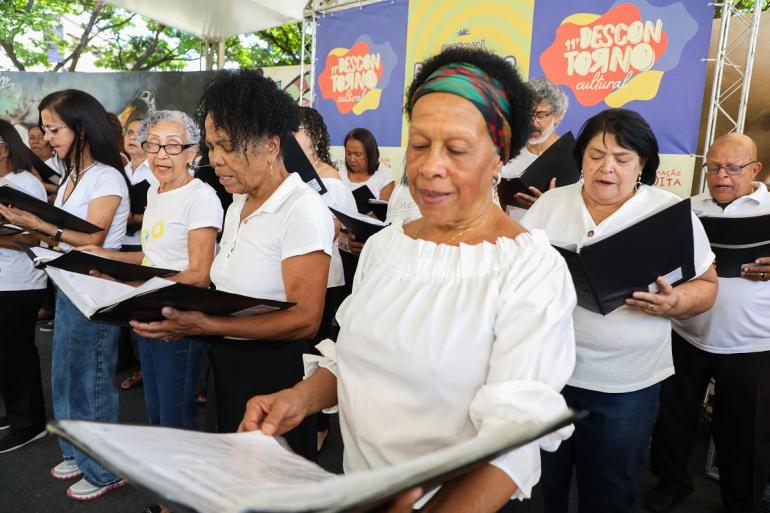 “Outubro Prateado” movimenta equipamentos culturais com programação para o público 60+ Apresentação do coral do projeto Saúde Encanto no Centro Cultural Urucuia. Em primeiro plano, uma mulher negra idosa veste blusa branca de ombro a ombro e faixa colorida no cabelo, segurando uma pasta preta de partitura aberta e cantando com os olhos direcionados para a folha. Ao lado e atrás dela, outros integrantes do coral, vestindo roupas brancas, acompanham a música com suas pastas. Ao fundo, há um painel colorido com a inscrição "11º Descontorno Cultural".