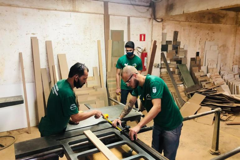 Três homens usando camisas verdes trabalham com máquinas e ferramentas em peças de marcenaria