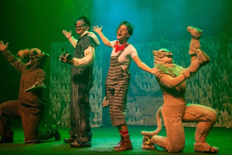 Teatro Francisco Nunes recebe espetáculo que une aventura e conscientização ambiental Cena do espetáculo "Margot e a Árvore da Vida" no palco, sob luzes verdes. No centro, uma mulher de camiseta branca, lenço vermelho no pescoço e jardineira listrada canta alegremente de braços abertos. Ao lado dela, um homem sorri usando macacão jeans, jaqueta de lã sintética e óculos de proteção. À esquerda, um ator ajoelhado veste uma fantasia marrom e uma máscara. À direita, outro ator ajoelhado usa uma fantasia felina, erguendo a perna e o braço.