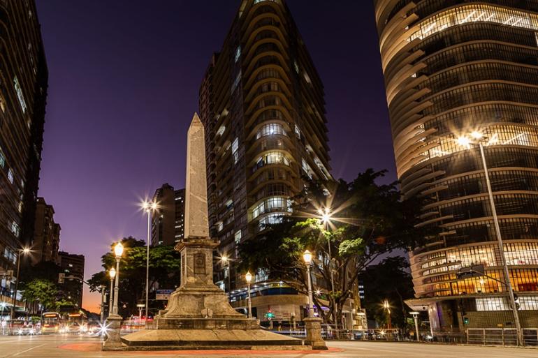 Plano geral noturno da Praça Sete de Setembro, no centro urbano de Belo Horizonte. No centro da cena, em primeiro plano, destaca-se o monumento do Obelisco — popularmente conhecido como "Pirulito da Praça Sete" —, iluminado por postes de luz ao seu redor. Ao fundo, altos edifícios iluminados emolduram a praça contra o céu em tom roxo-escuro do anoitecer.