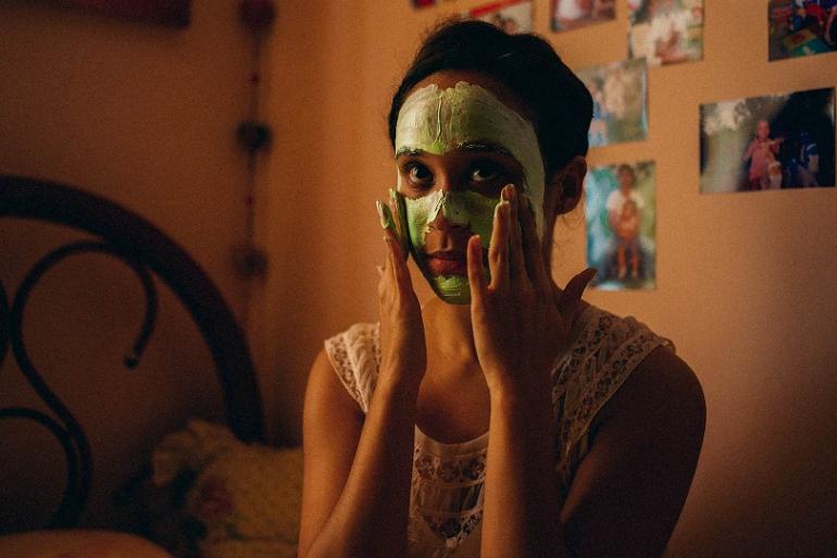 Jovem mulher com o rosto coberto por uma máscara facial verde aplicando o produto com as mãos, em cena do filme Medusa. Ela está em um quarto aconchegante com iluminação alaranjada e fotos na parede ao fundo.