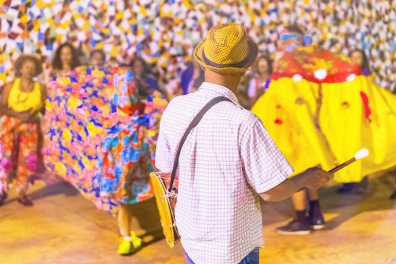 Memórias, saberes e tradições ganham destaque na programação do Mês do Patrimônio Cultural Fotografia colorida em plano médio de uma apresentação cultural festiva. Em primeiro plano, centralizado e de costas para a câmera, um homem vestindo camisa xadrez clara, chapéu de palha e calça jeans segura uma baqueta com a mão direita e carrega um tambor a tiracolo.